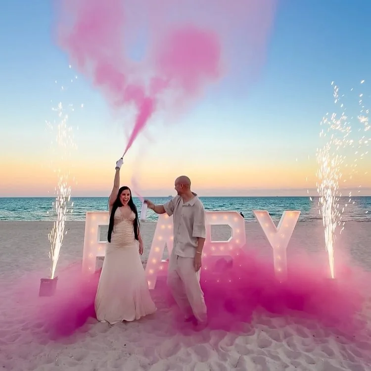 Couple celebrating a pink powder gender reveal beside illuminated BABY letters at sunset
