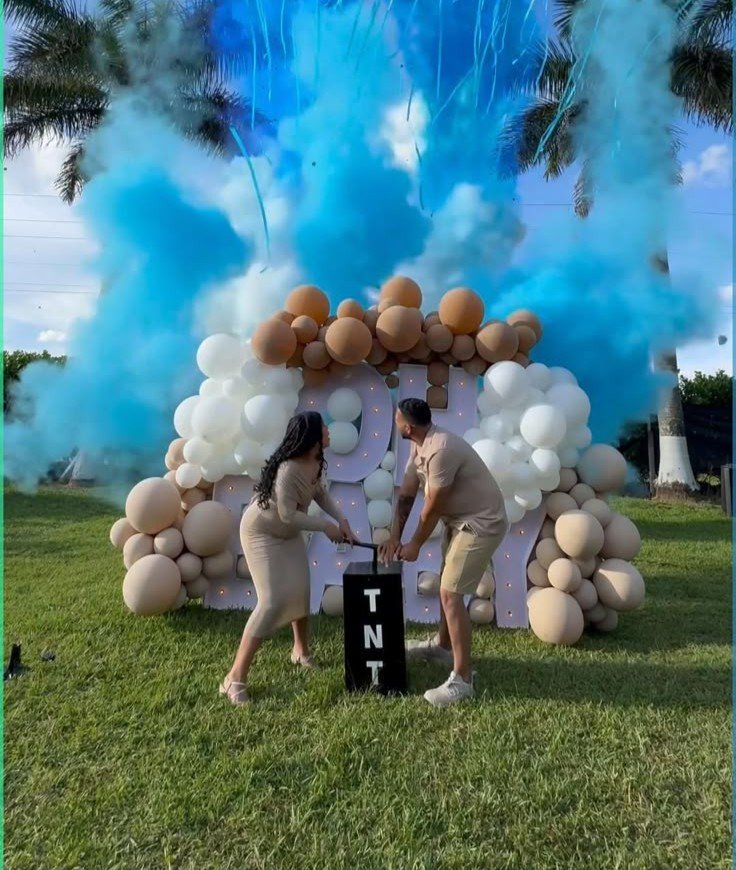 Couple beside a TNT box, balloon backdrop and blue powder cloud at an outdoor gender reveal